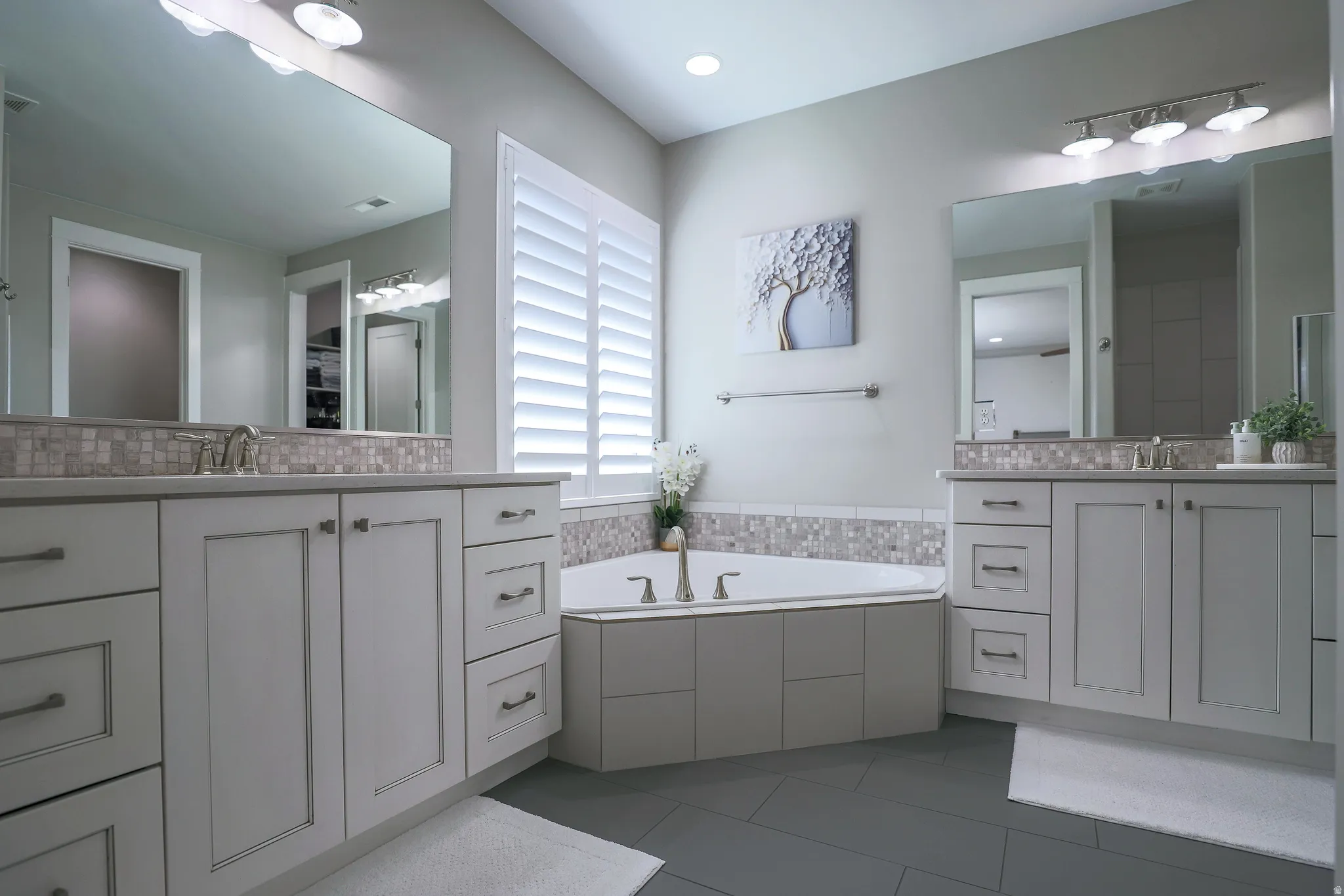 Modern bathroom featuring a built-in corner soaking tub, dual vanities with light gray cabinetry, and a mosaic tile backsplash