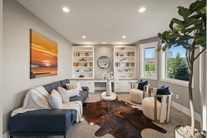 Recessed lighting illuminates a versatile room featuring a built-in desk with white cabinetry, open shelving, and two large windows offering natural light