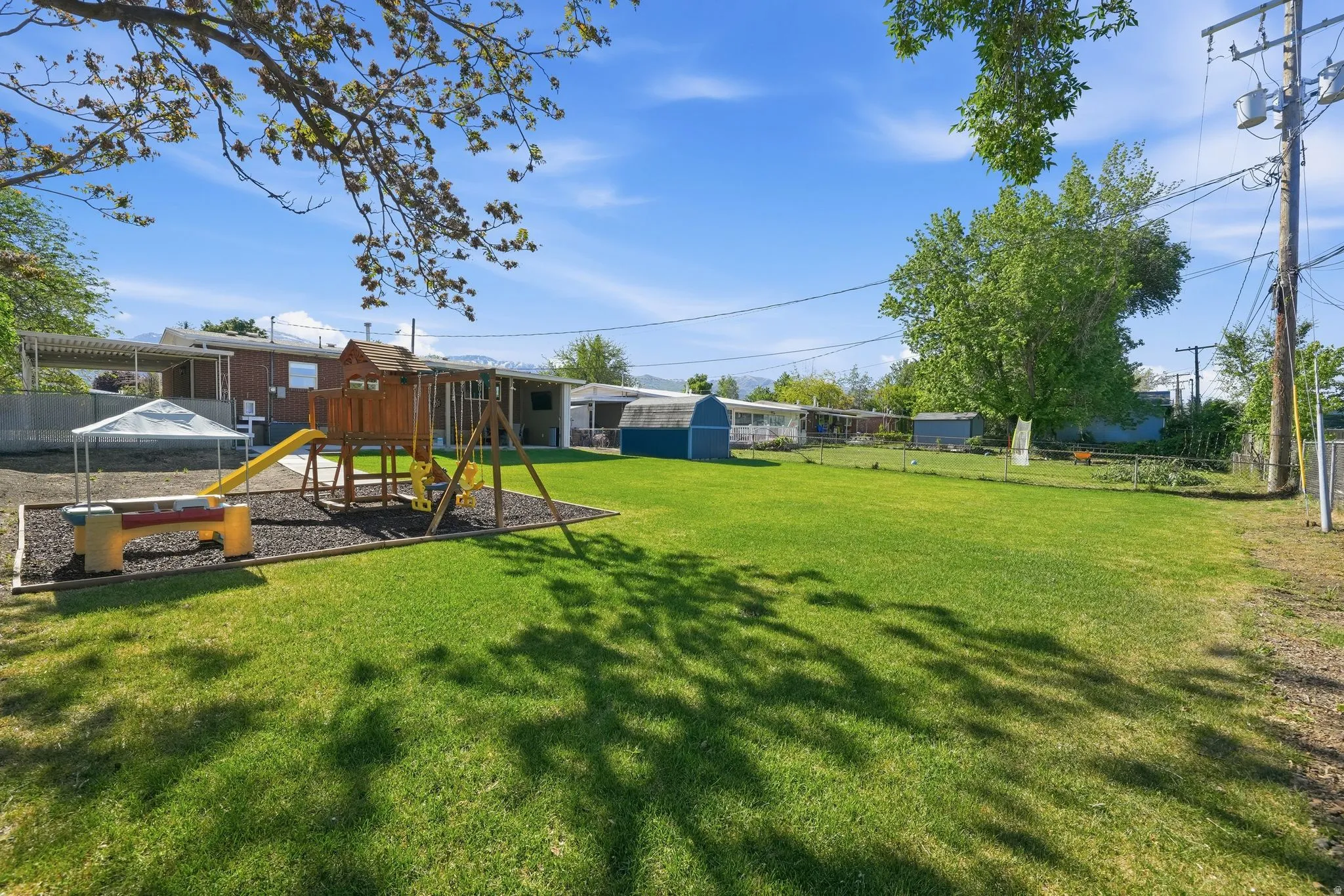 Expansive grassy yard area featuring a built-in wooden playset with slide and swings, a covered picnic table, and mature trees