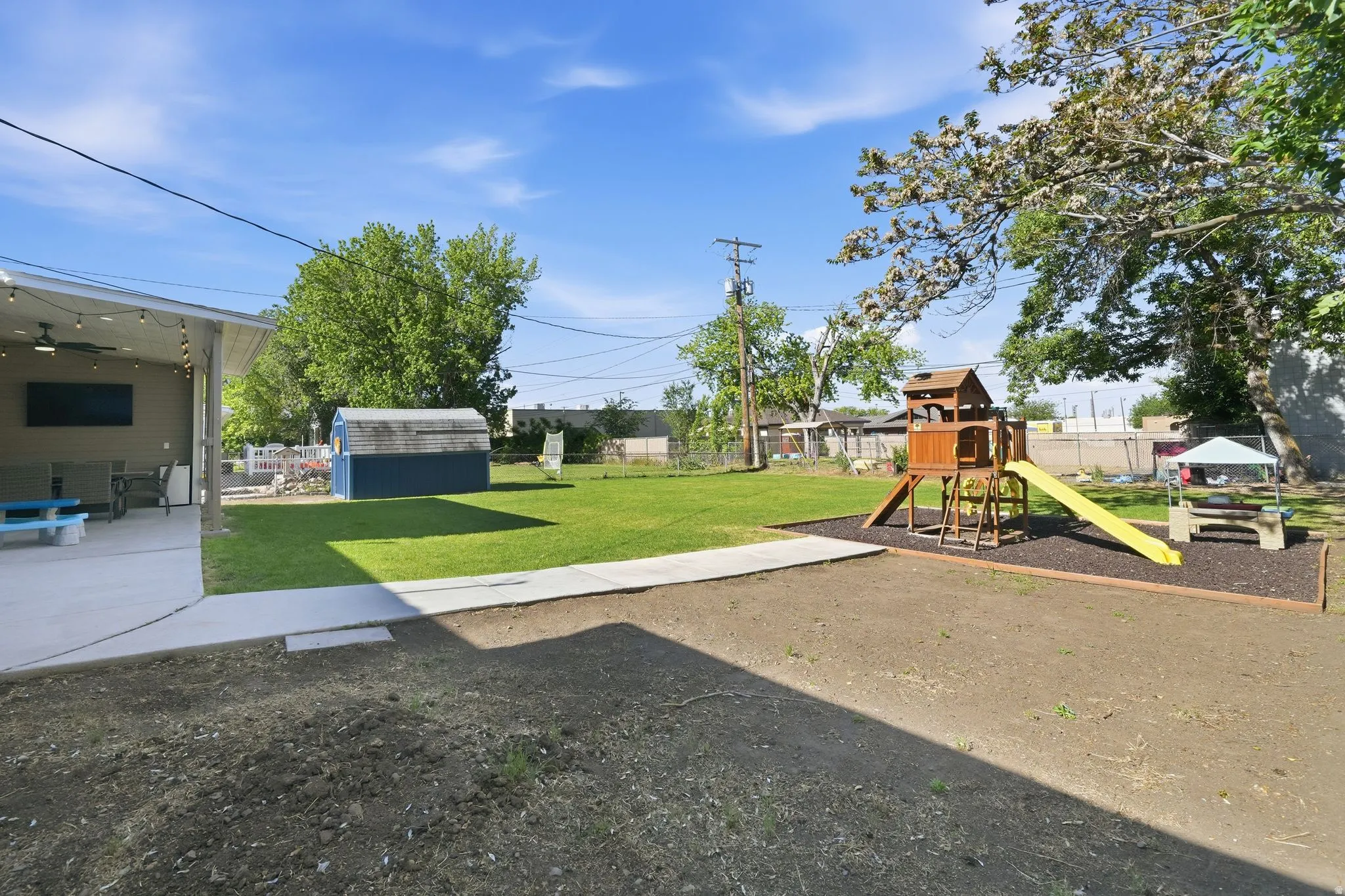 Expansive backyard featuring a covered patio with string lighting and ceiling fan, a detached storage shed, and a dedicated play area with a wooden structure and slide