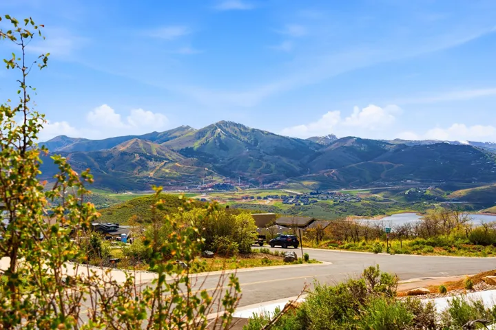 Expansive mountain and valley scenery featuring a prominent body of water, distant residential structures, and a winding paved road