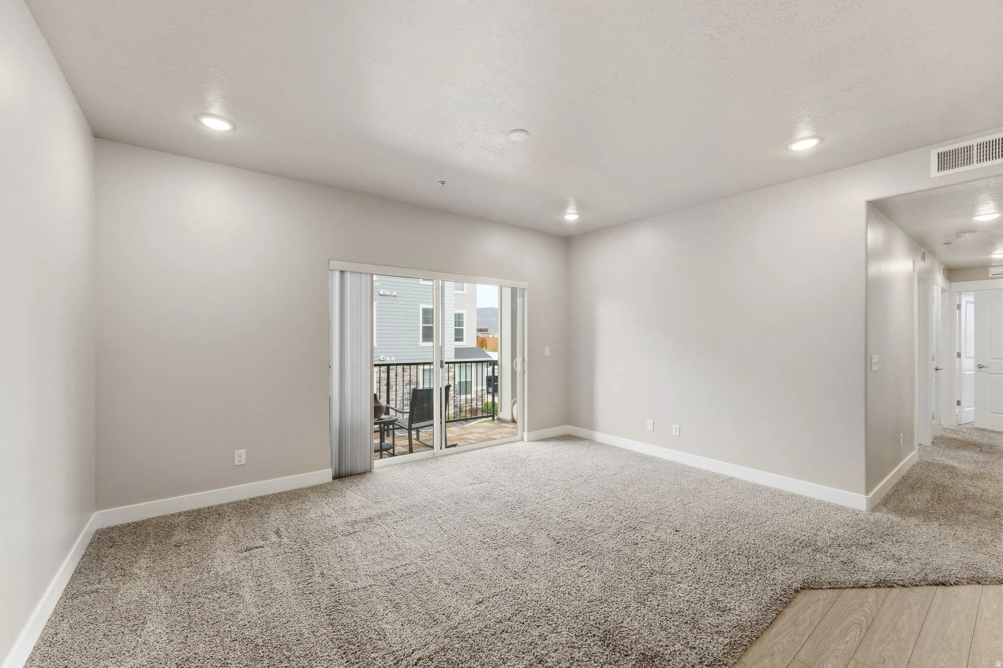 Spacious living area featuring neutral carpeting, recessed ceiling lighting, a sliding glass door to a private balcony, and light-toned walls