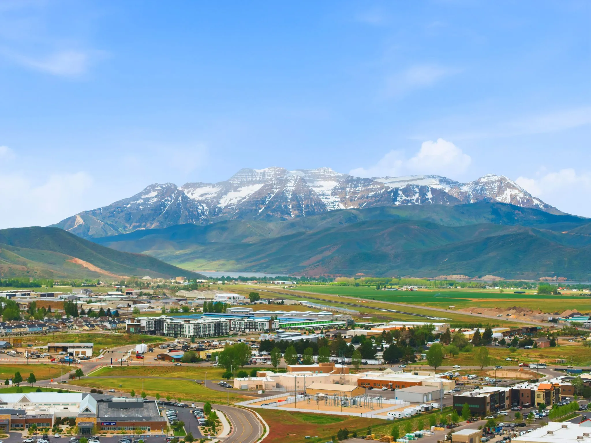Expansive landscape featuring snow-capped mountain peaks, a body of water, green fields, and a developing community