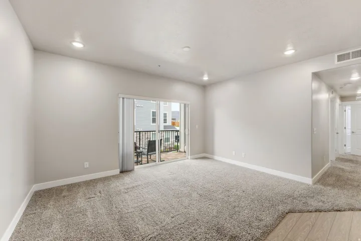 Spacious living area featuring neutral carpeting, recessed ceiling lighting, a sliding glass door to a private balcony, and light-toned walls