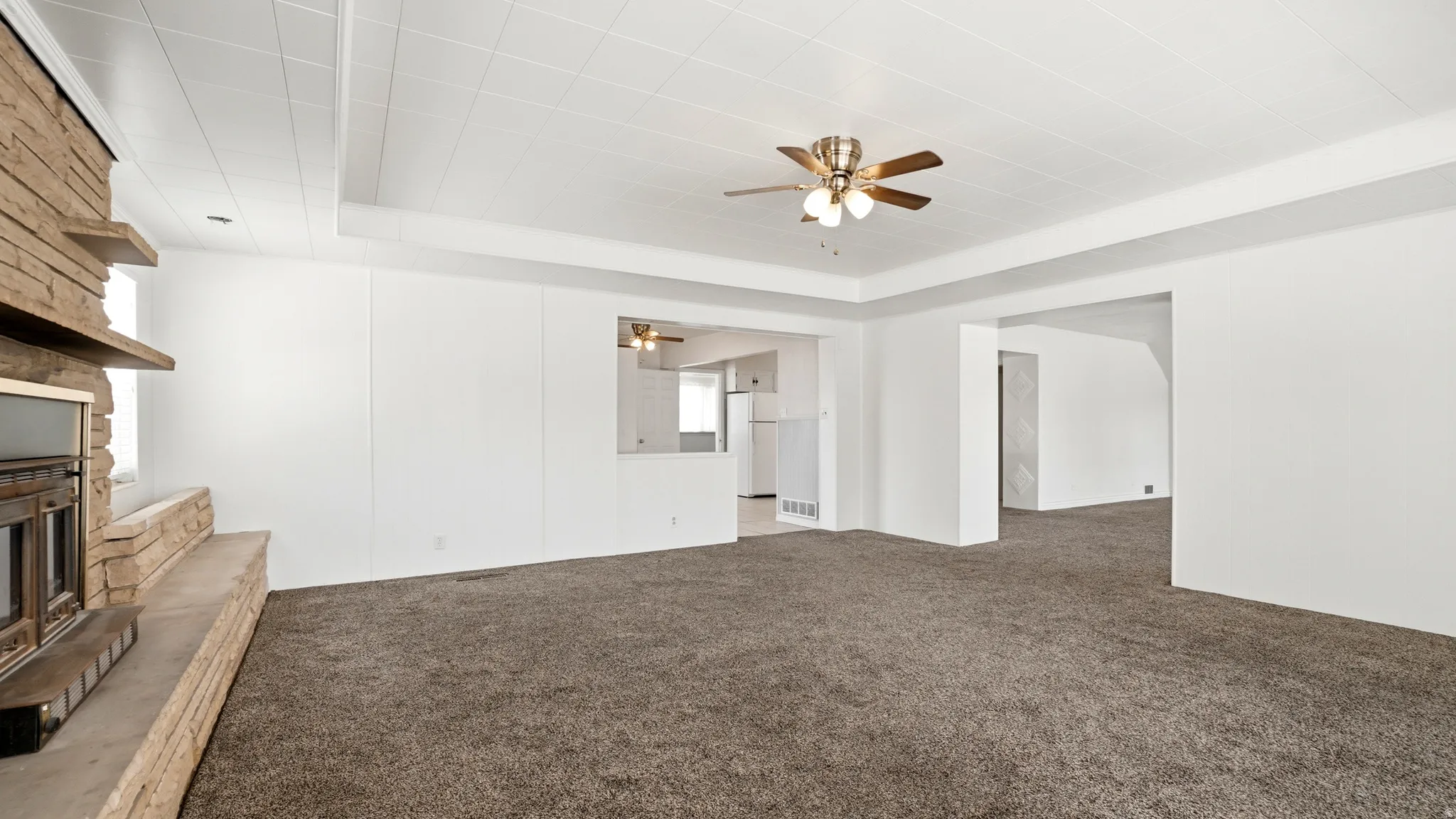 Spacious room featuring a stone fireplace with a hearth, wall-to-wall carpeting, white walls, a ceiling fan with light fixture, and a pass-through window to an additional area
