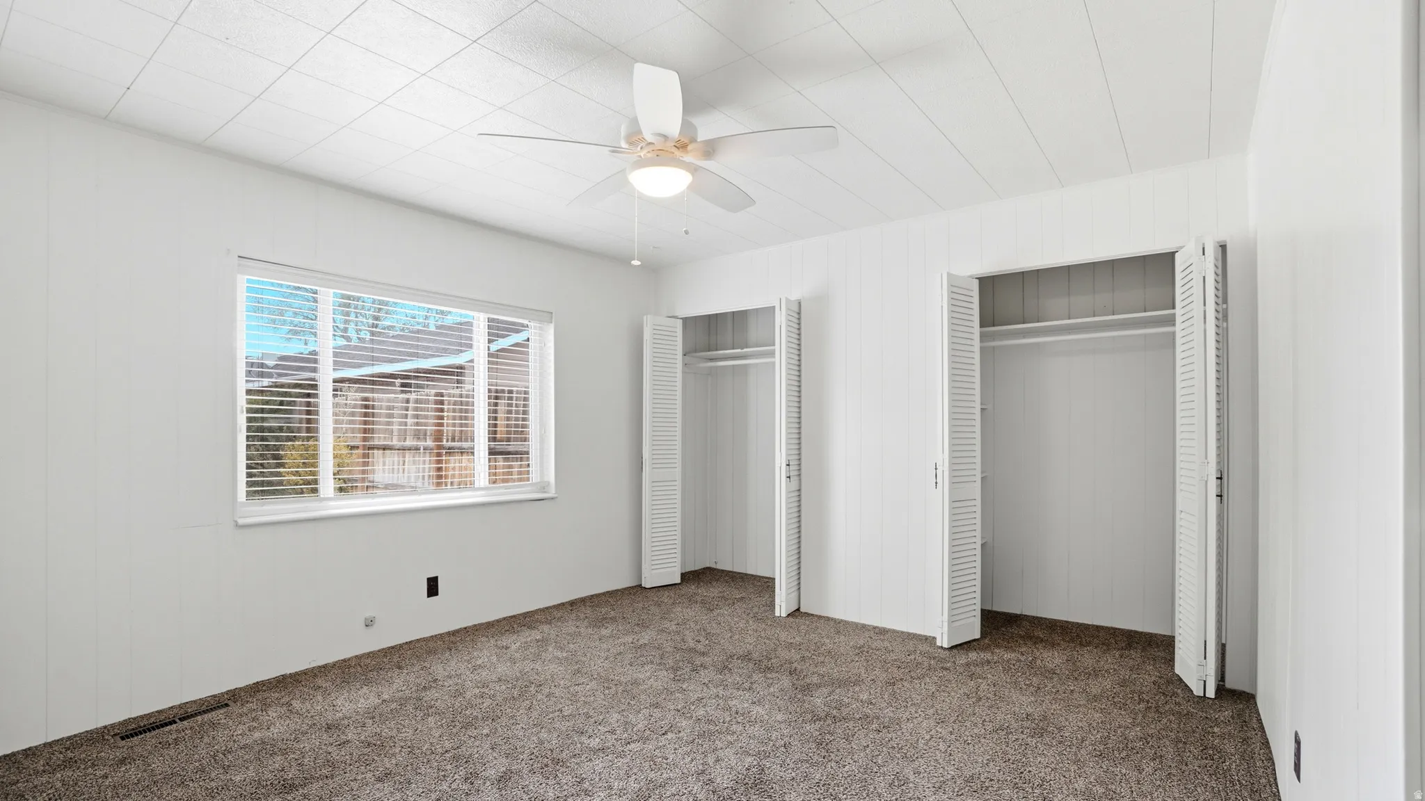 Carpeted room featuring a white ceiling fan with integrated lighting, a wide window with blinds, and two closets with louvered bifold doors