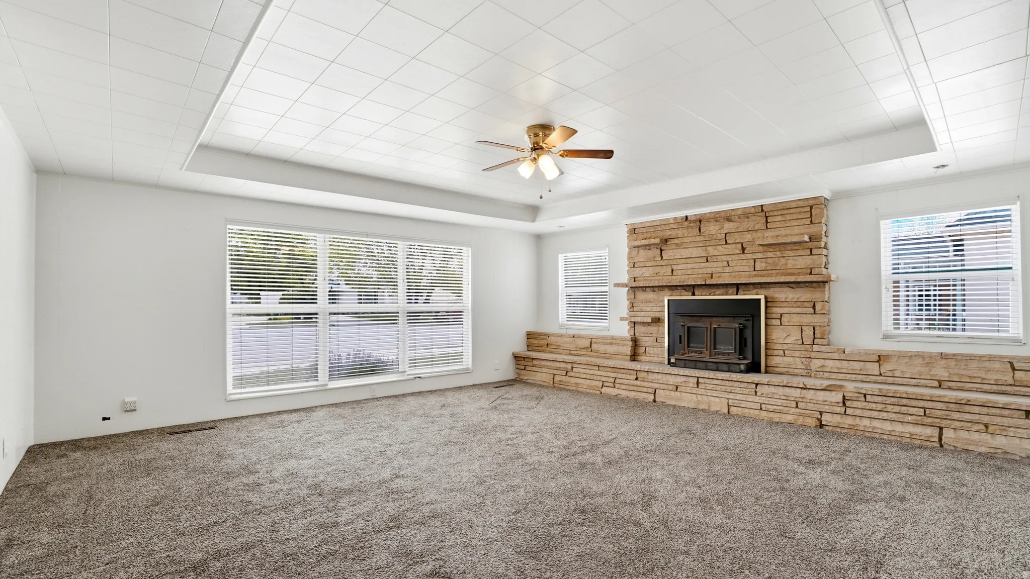 Spacious living area featuring a prominent stone-clad fireplace with a black insert, large picture windows, recessed lighting, a ceiling fan, and grey carpet flooring