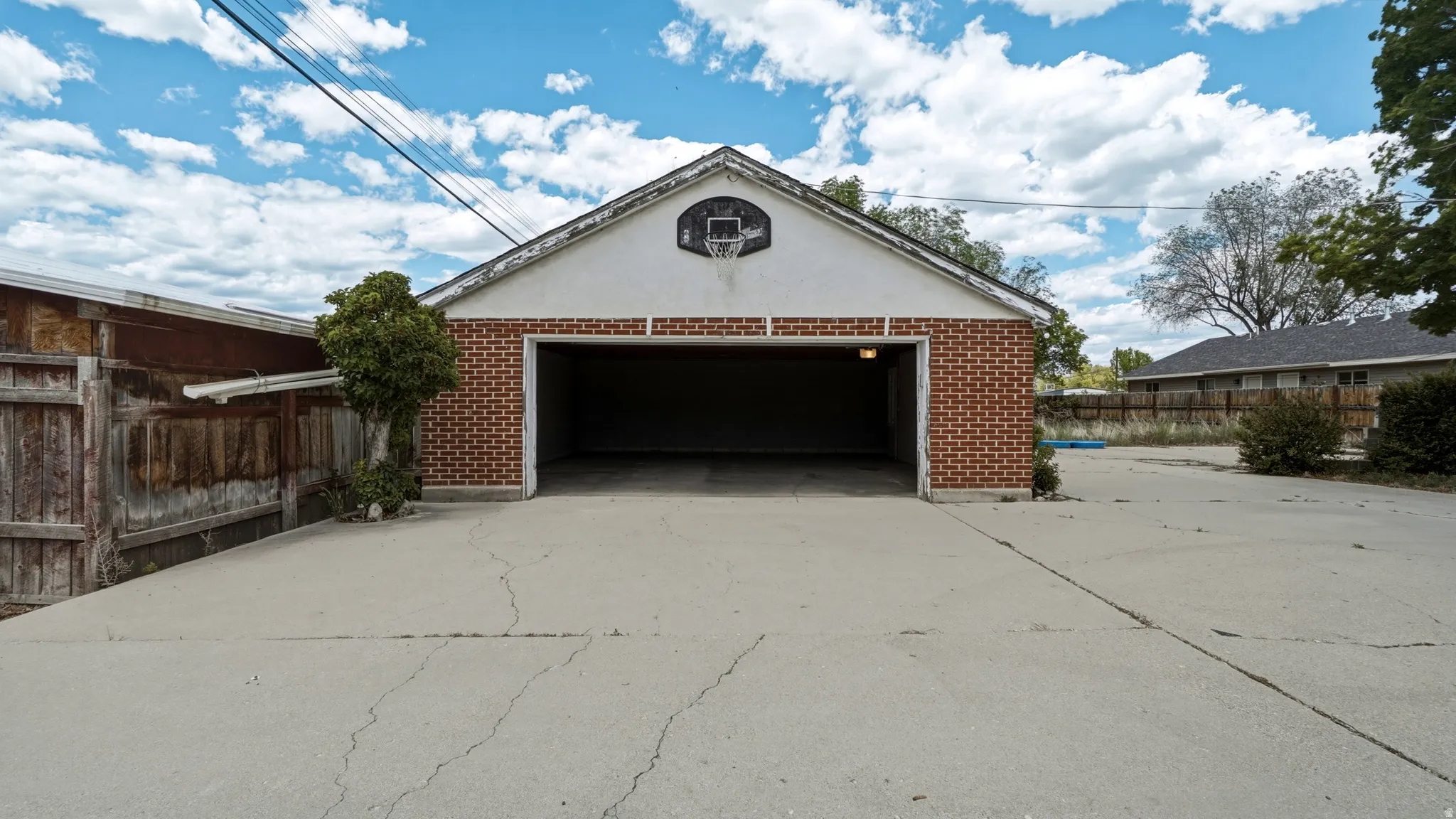 Detached brick garage with an exposed beam ceiling and concrete flooring, featuring an open entry and an exterior light fixture