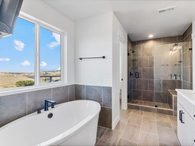 Standalone soaking tub with black fixtures, expansive window, tiled shower with glass enclosure and built-in bench, gray large-format tile flooring, and a white vanity with black hardware