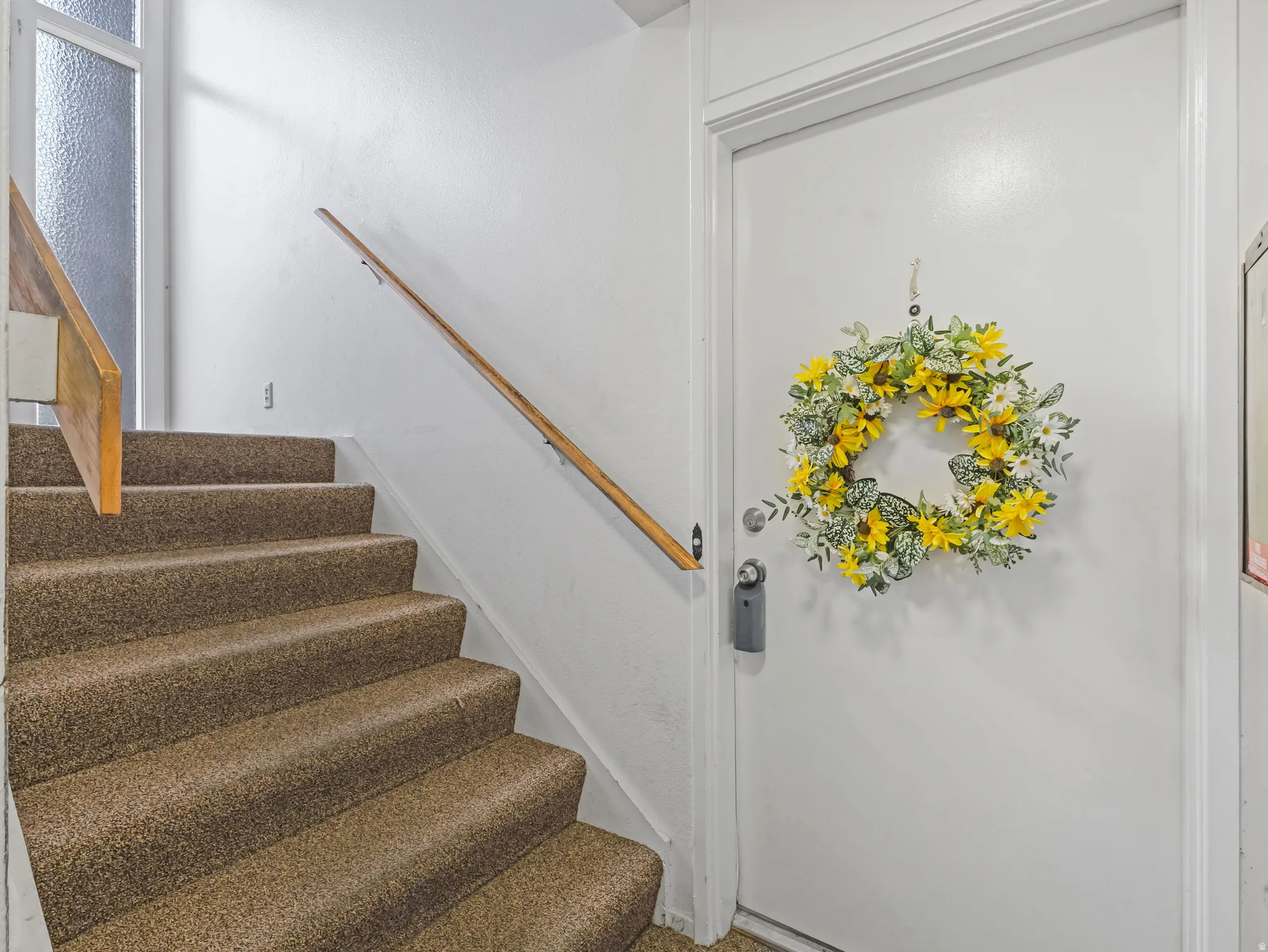 Carpeted staircase with a wood-finish handrail, white painted walls, and a white entry door with a privacy lock