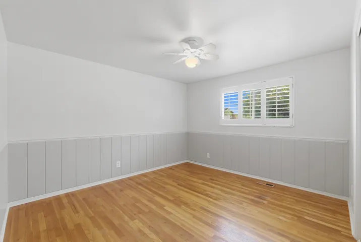 Bright room featuring wood-finish flooring, half-wall paneling, a ceiling fan with integrated lighting, and a window with plantation shutters
