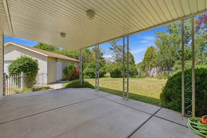 Covered concrete patio with decorative metal support posts, offering views of a spacious lawn and mature landscaping