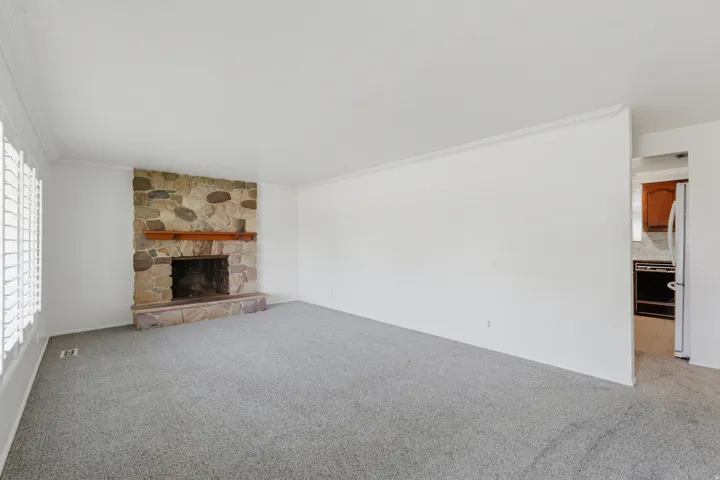 Spacious living area featuring a stone fireplace with a wood mantle, neutral carpeting, and white plantation shutters