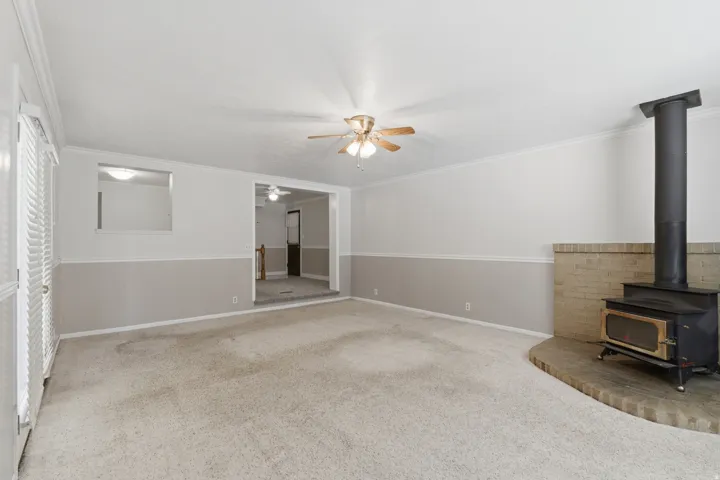 Spacious carpeted room featuring a wood-burning stove with a brick hearth, a ceiling fan with integrated lighting, and a two-tone wall finish with wainscoting