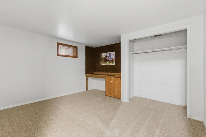 Carpeted room featuring a built-in desk with wooden cabinetry, a wood-framed window with blinds, and an open closet