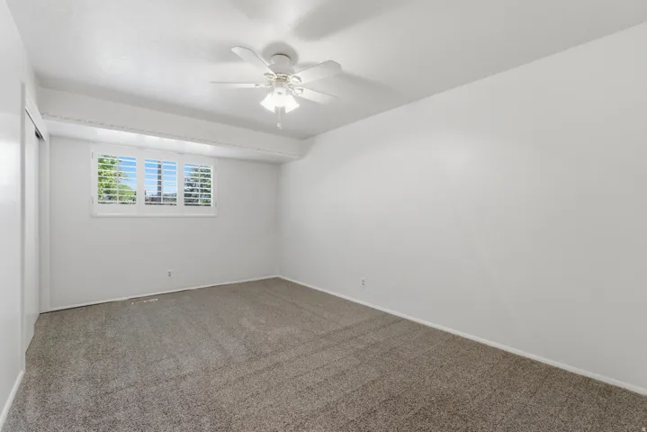 Carpeted room featuring a white ceiling fan with integrated lighting, a window with white plantation shutters, and a sliding door closet