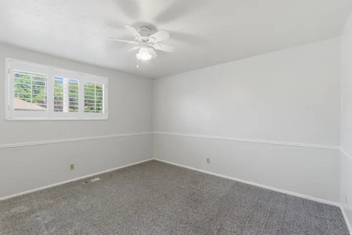 Neutral interior room featuring white window shutters, a ceiling fan with integrated lighting, and gray carpeting