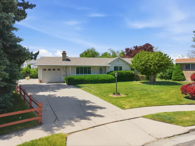 Ranch-style residence featuring a single-car garage with arched window accents, a prominent front lawn with mature trees and shrubbery, and a concrete driveway leading to the street