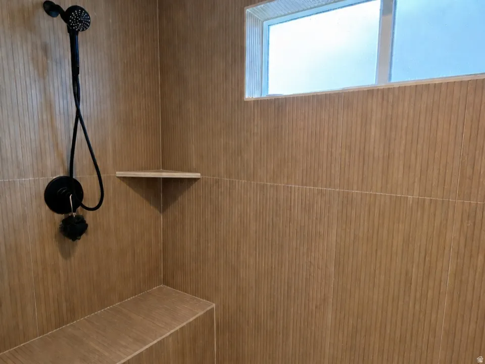 Primary ensuite walk-in shower featuring patterned tile flooring, wood-look wall tile, dual shower heads, a built-in shower bench, a corner shelf, and a frosted transom window