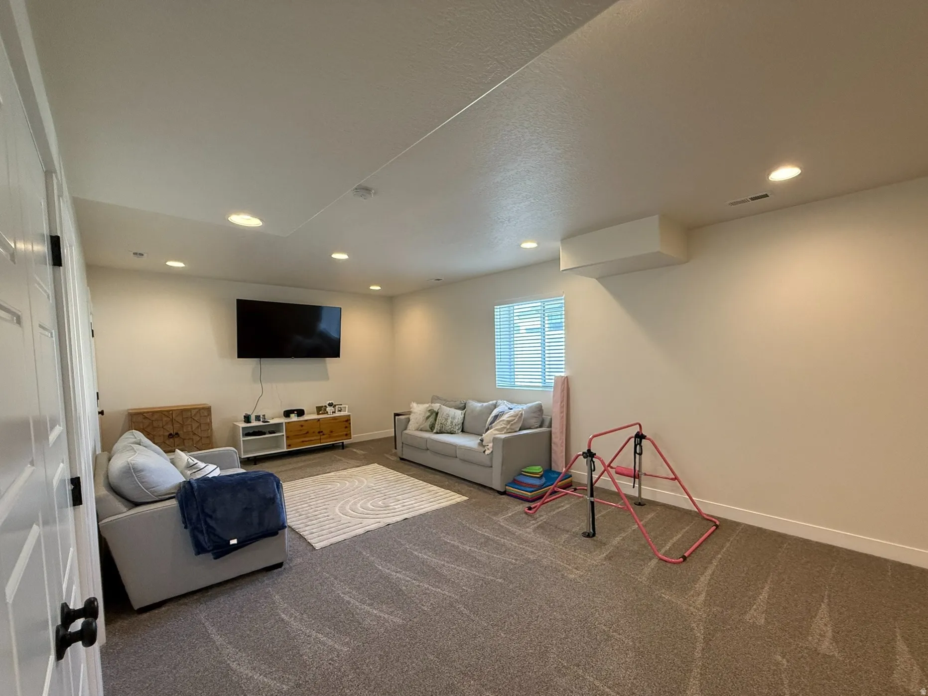 Spacious family room featuring neutral carpeting, recessed ceiling lighting, a wall-mounted television, and a window with blinds