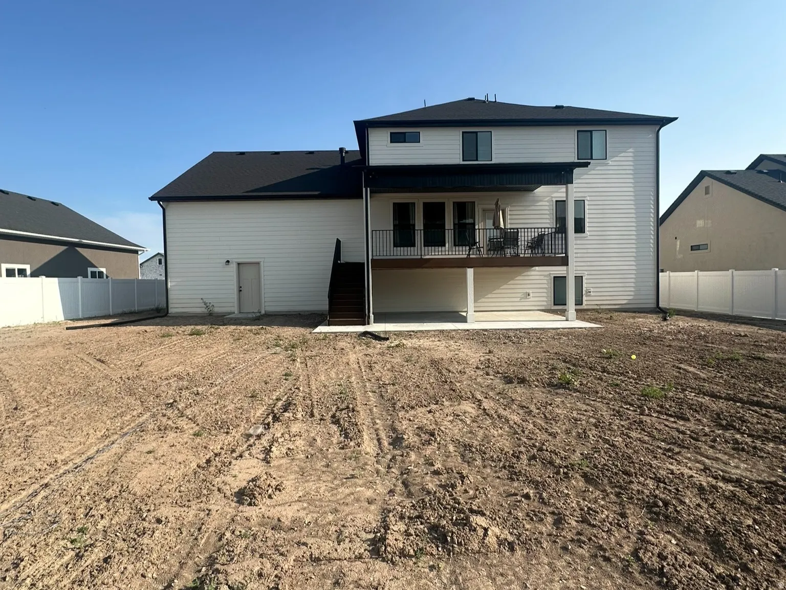 Rear exterior featuring a second-story deck with black metal railings, a ground-level concrete patio, and a spacious undeveloped and partially fenced yard