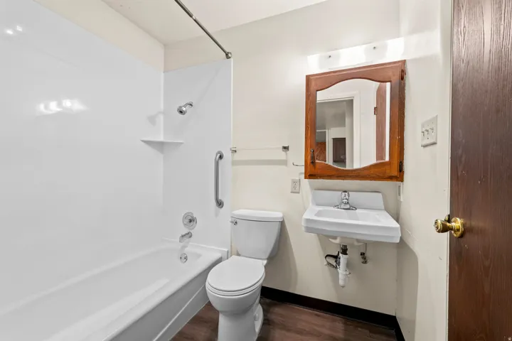 Bathroom featuring a white tub and shower surround, a wall-mounted sink, a toilet, and wood-finish flooring