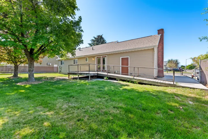 Rear exterior featuring a spacious lawn, mature trees, a wooden deck with railing, a ramp for accessibility, and a brick chimney
