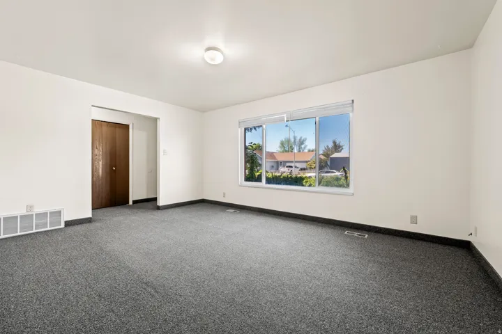 Spacious room featuring neutral white walls, dark gray carpeting, and a large window providing natural illumination
