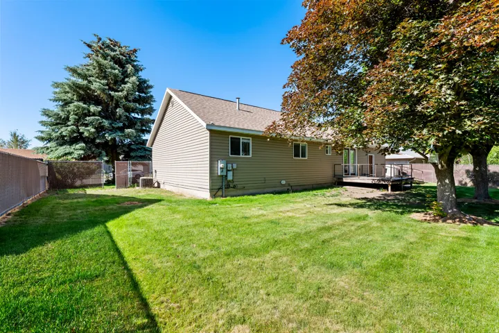 Spacious grassy backyard featuring mature trees providing shade, a perimeter chain-link fence, and a wooden deck