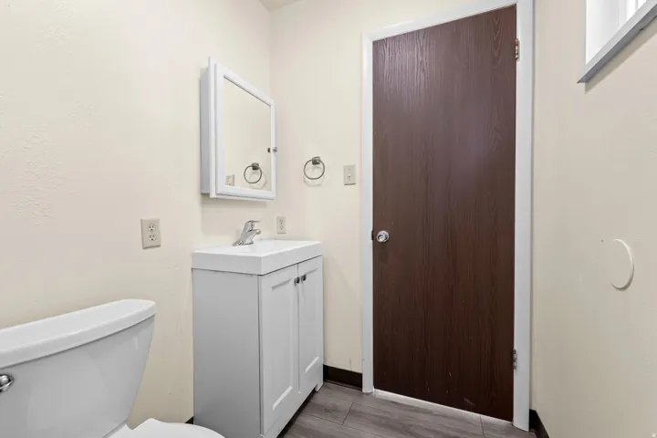 Bathroom featuring a white toilet, a white vanity with an integrated sink, a mirrored medicine cabinet, and wood-finish flooring