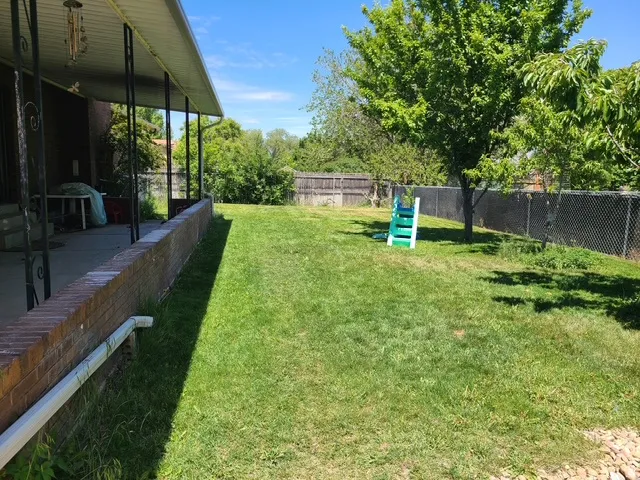 Spacious backyard featuring a chain-link fence, mature trees, and a brick retaining wall bordering a covered patio