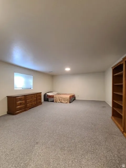 Spacious room featuring light gray carpeting, recessed ceiling lighting, a window with horizontal blinds, and a built-in wood bookshelf with multiple shelves