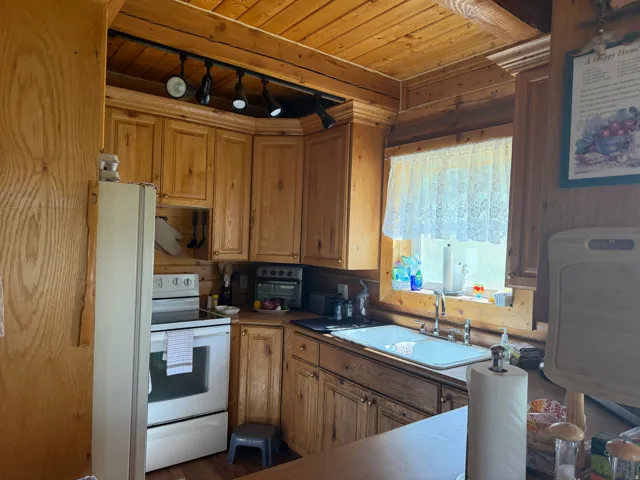 Kitchen featuring wood-paneled walls and ceiling, natural wood cabinetry with brass-toned hardware, a white double basin sink with a chrome faucet, a white electric range, and a rectangular window with lace curtains