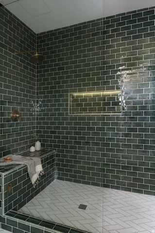 Spacious walk-in shower featuring dark green subway tile walls, a built-in bench, a recessed niche with integrated lighting, and white herringbone tile flooring