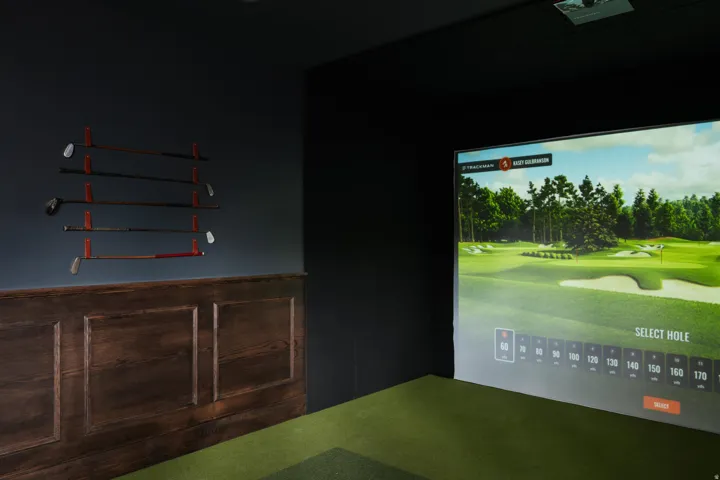Dedicated golf simulator room featuring a large projection screen, dark wall finishes, and a green turf floor