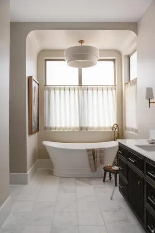 Freestanding soaking tub set within a windowed alcove, featuring a tiered drum chandelier, white tiled flooring, and a dark wood vanity with a white countertop