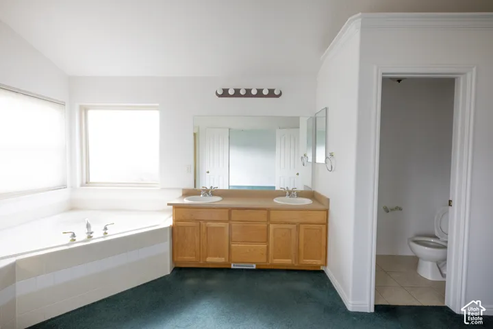 Corner soaking tub with tiled surround, dual-sink vanity with wood cabinetry, and a separate toilet area with tiled flooring