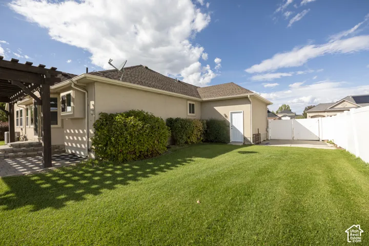 Expansive green lawn, stucco exterior, rear patio with a wooden pergola, privacy fence, and a paved side access area