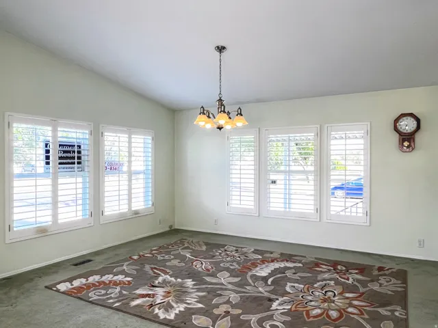Vaulted ceiling with a central chandelier, multiple windows with plantation shutters, light-toned walls, and carpet flooring