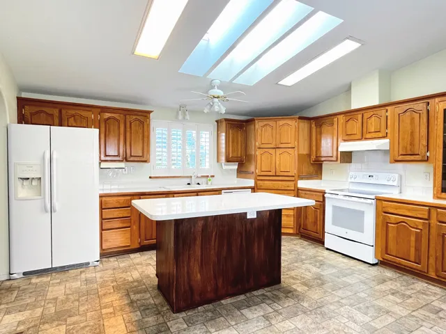 Kitchen featuring a central island with a light-toned countertop, wood cabinetry, white appliances, and a white subway tile backsplash