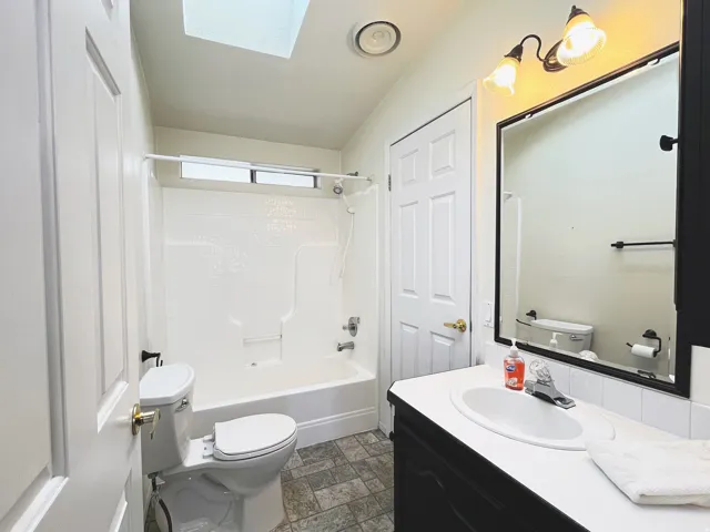 Full bathroom featuring a white tub/shower combination with a transom window, a white toilet, and a vanity with a white countertop and dark base