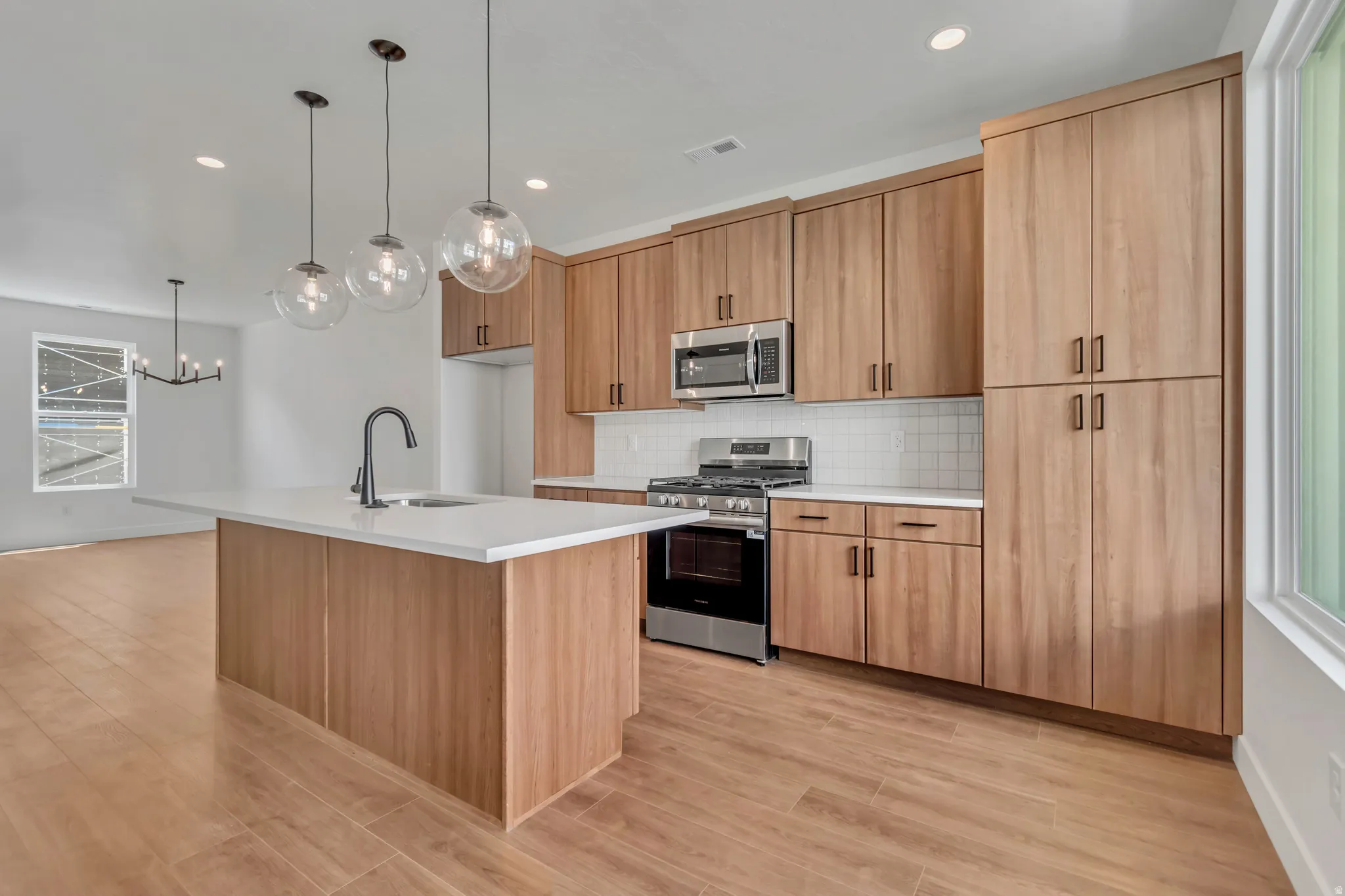 Kitchen featuring a central island with a sink and matte black faucet, pendant lighting, wood-finish cabinetry, stainless steel appliances, and wood-finish flooring