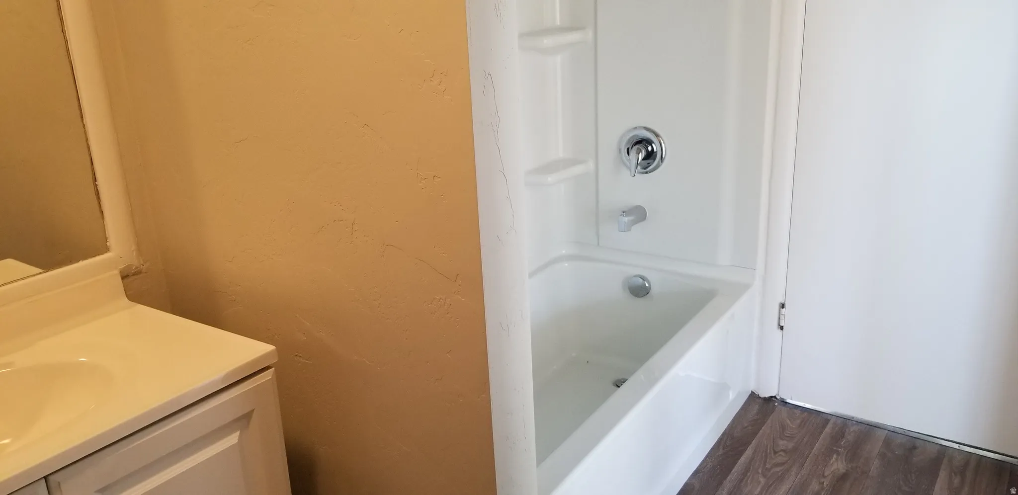 White fiberglass tub and shower combination with chrome fixtures, wood-finish flooring, and a single-sink vanity with white cabinetry and a matching countertop