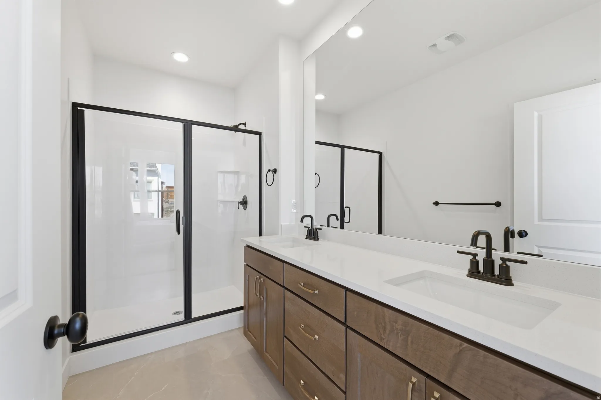 Bathroom featuring a dual vanity with a white countertop and bronze-finish fixtures, wood-finish cabinetry, a large frameless mirror, a walk-in shower with black-framed glass doors, and recessed lighting