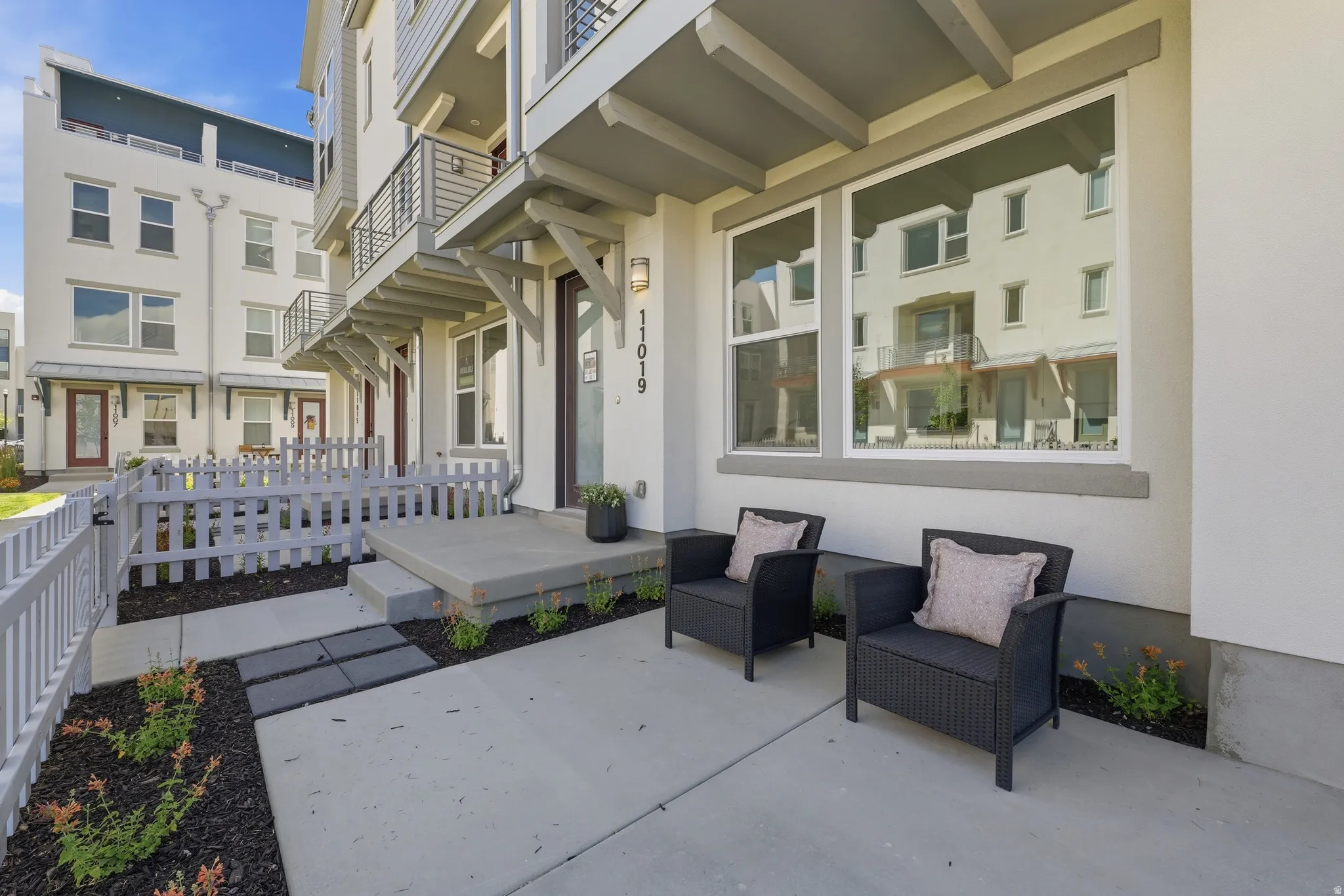 Contemporary townhome exterior featuring a private fenced front yard, concrete patio, modern wall sconce lighting, and a white picket fence