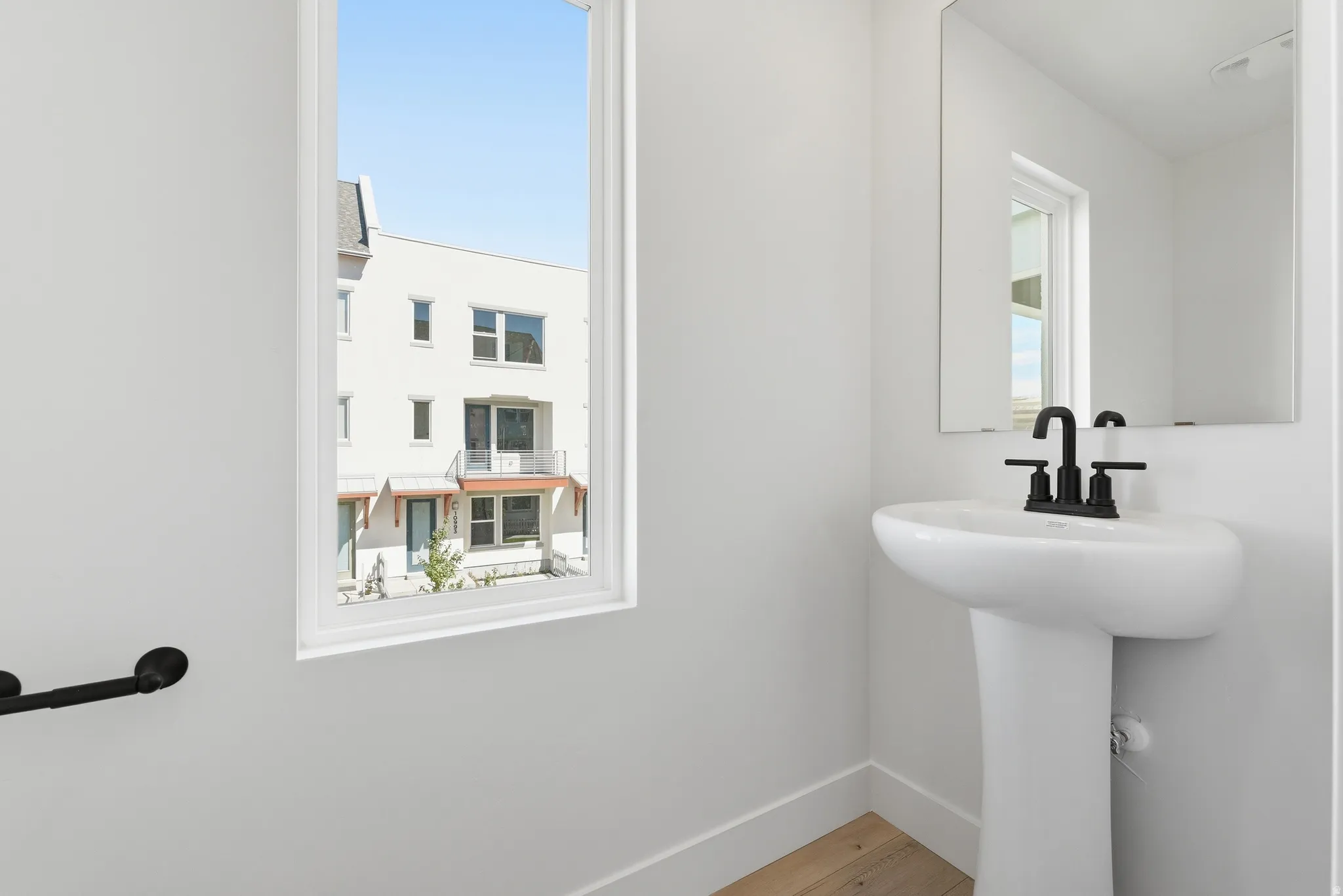 Pedestal sink with matte black faucet, wall-mounted mirror, and wood-finish flooring