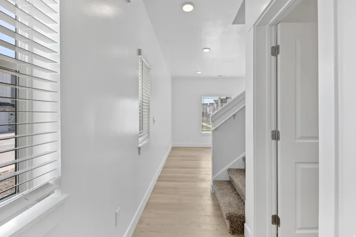 Hallway with light wood-finish flooring, white walls, recessed lighting, and windows featuring horizontal blinds