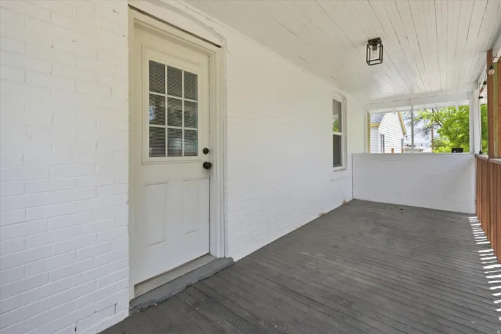 Covered back porch featuring a painted brick exterior, wood-finish flooring, a beadboard ceiling, a multi-pane glass door, and a modern ceiling fixture