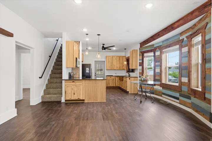 Open-concept living area featuring rich wood-finish LVP flooring, a kitchen with light wood cabinetry and dark countertops, a decorative accent wall with varied wood paneling, and a staircase with a dark handrail