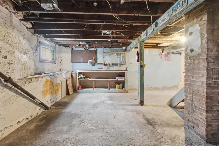 Unfinished basement featuring exposed wood joists, concrete flooring, a brick support column, a small window, and built-in shelving